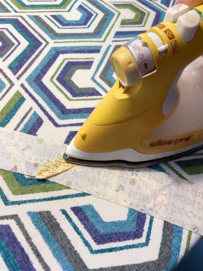 A yellow Oliso Pro iron is shown pressing the diagonal seam between two yellow strips of fabric open. The background is a white, teal, blue and gray geometric ironing board cover. 