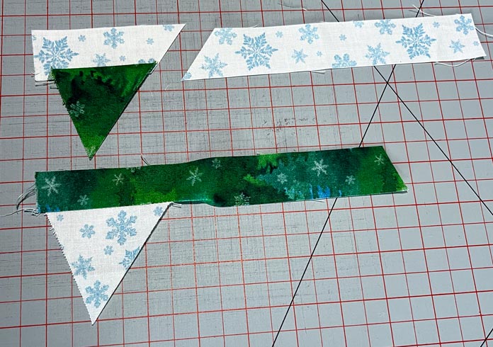 A strip of fabric is sewn to a triangle piece of fabric, pressed, and trimmed; another strip is sewn to another triangle piece of fabric, pressed but not trimmed. Fabric is white with blue snowflakes and dark green; Heirloom 24″ x 36″ Double Sided Cutting Mat