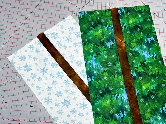 A brown strip sewn between two snowflake and two green fabric strips lay on top of a gray cutting mat with red gridlines