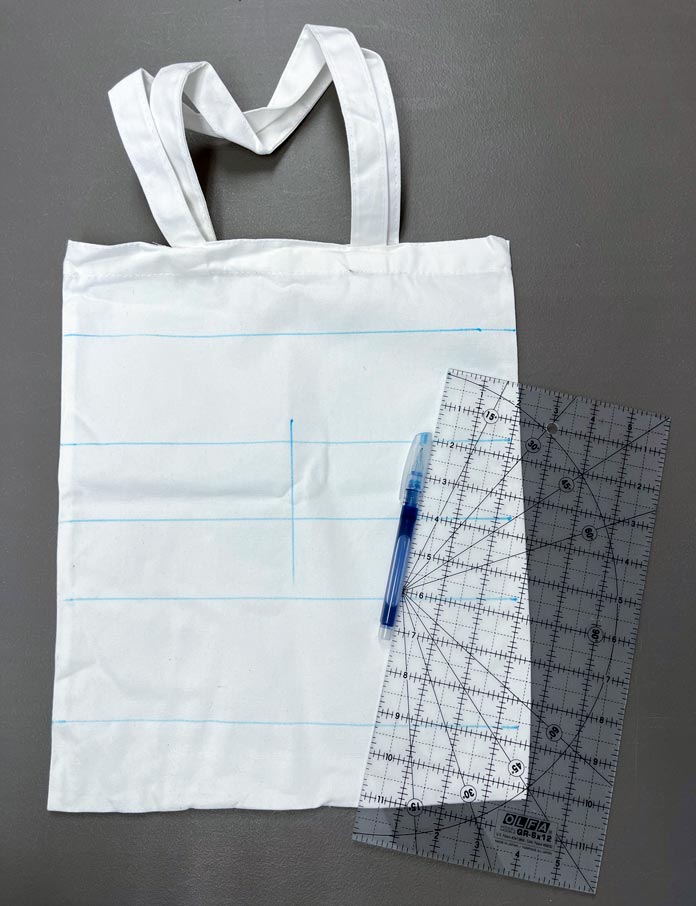 A light colored canvas tote bag marked with blue lines next to a blue wash out marker and OLFA 6” x 12” quilting ruler