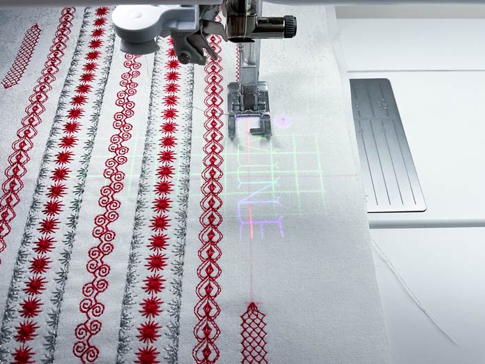 Using the projector on my PFAFF creative icon 2 to position and stitch the text between the tapered stitches in red and metallic threads on white fabric 