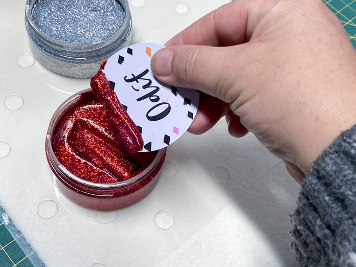 A hand is shown holding a white plastic applicator which has been dipped into a jar of red Odif OdiShine Glitter Gel. There is red glitter gel on the straight edge of the applicator. In the background, there is a clear plastic stencil stuck to a rectangle of cream-colored felted wool. The wool sits on green cutting mat.
