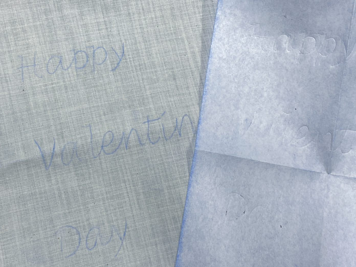 The words Happy Valentine’s Day show up on in blue markings on a cream-colored fabric. The back of a piece of blue transfer paper sits on top of the cream fabric.