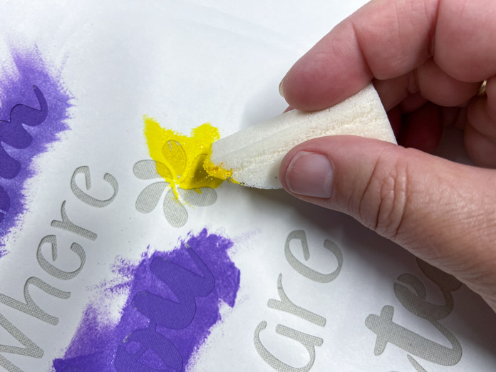 A hand holding a white triangular makeup sponge is shown applying yellow paint to the white stencil; Sew Easy Freezer Paper for Quilting and Applique, Mont Marte Signature Fabric Paints