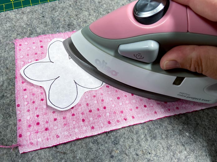 A pink Oliso Mini Project iron is ironing a white flower shape onto the back of a pink fabric. A gray wool pressing mat is shown in the background; UNIQUE Quilting Wool Pressing Mat - 14″ x 14″, HeatnBond EZ Print Feather Lite 10 pcs - 22 x 28cm (8½″ x 11″)