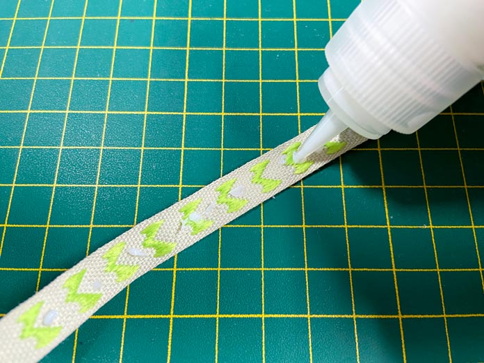 The tip of a small white bottle is shown placing small dots of adhesive onto the back of a green and white ribbon which is laying on a green cutting mat; Unique Creativ Trim Pack – Red and Green, Fabric Fuse by HeatnBond Quick Bond Fabric Adhesive