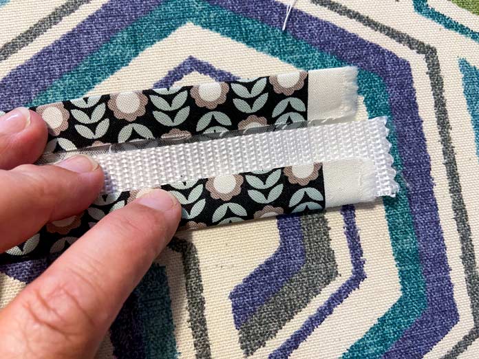 A hand is shown holding a folded strip of fabric that has a piece of white webbing inside. There’s a multi-colored ironing board cover in the background.