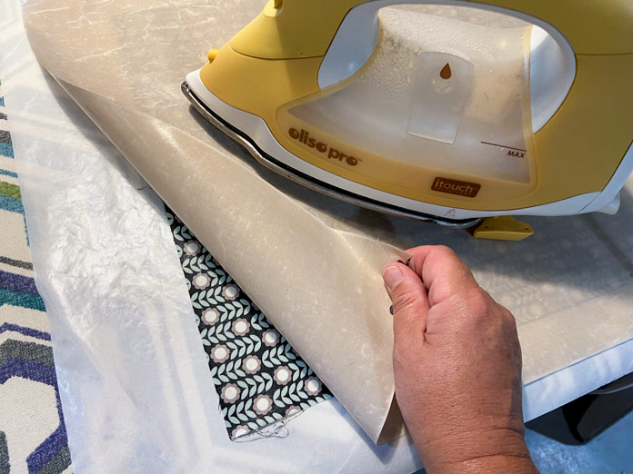 A hand folds back the corner of a brown applique pressing sheet to show a square of treated multicolored fabric. A yellow iron sits on top of the pressing sheet.