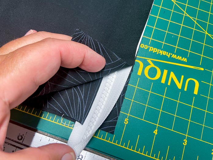 A hand is shown folding back the corner of a black fabric rectangle to show a zipper and another black fabric underneath. There is a green cutting board in the background.