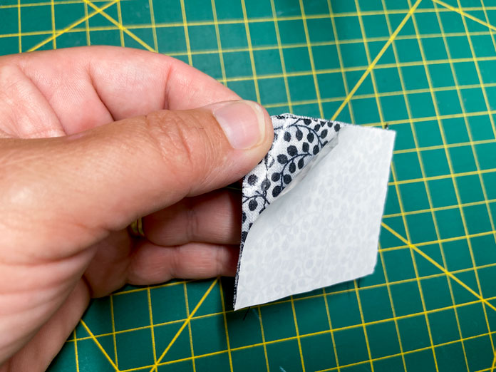 A hand peels back the paper backing of a rectangle of HeatnBond EZ PRINT FEATHERLITE to show the adhesive stuck to a black and white fabric pouch. A green cutting mat is shown in the background.