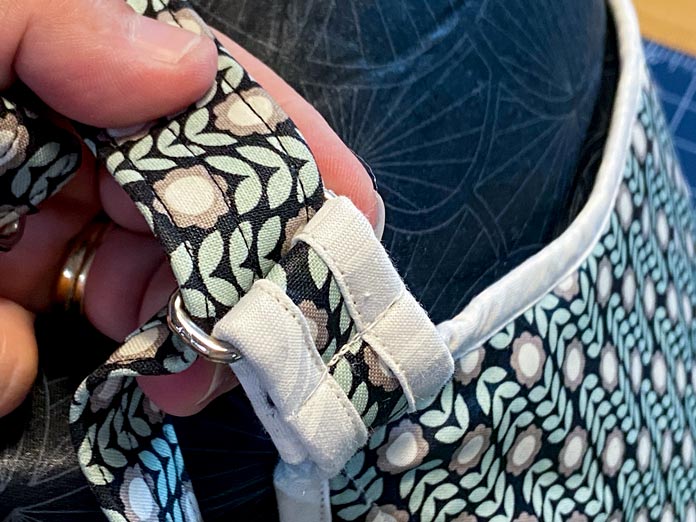 A hand is shown threading a multicolored fabric strap through a silver rectangular ring on a crossbody bag.