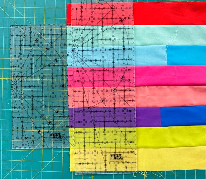 Two OLFA rulers are placed side by side to create a 6½” measurement on top of a strip set of 8 brightly colored fabrics on a green cutting mat.