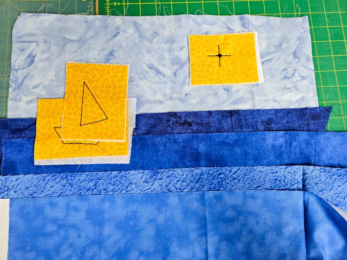 Yellow fabric squares on strips of blue fabric with a green cutting mat in the background. Husqvarna VIKING DESIGNER EPIC 3 sewing and embroidery machine, Husqvarna VIKING 6in/15.2cm Applique Scissor, Husqvarna VIKING Open Toe Presser Foot for IDF System, SULKY Slimline Metallic Dream Assortment