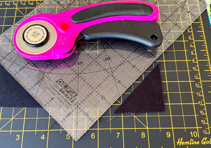 A pink OLFA cutter and frosted OLFA ruler are being used to trim a 45-degree corner from two strips of black fabric on top of a green cutting mat. Hemline Gold Multi-Use Craft Bag (28.5cm x 36.5cm x 17.8cm), Hemline Gold Sewing Kit, Hemline Gold Quilters 4-in-1 Multi-Mat (30cm x 24cm), Hemline Gold Quilters Clips (Pack of 30), Hemline Gold Retractable Tape Measure - 150cm/60, oliso M3Pro Project Iron – Coral, OLFA 45mm RTY-2/DX/MAG Ergonomic Rotary Cutter – Magenta, OLFA RM-MG - 24″ x 36″ Double Sided Rotary Mat, UNIQUE Sewing Tools Holder, OLFA QR-6x12 - 6″ x 12″ Frosted Acrylic Ruler, OLFA QR-6x24 - 6″ x 24″ Frosted Acrylic Ruler, Fairfield Quilter's 80/20 Quilt Batting - 229 x 274cm (90″ x 108″) 