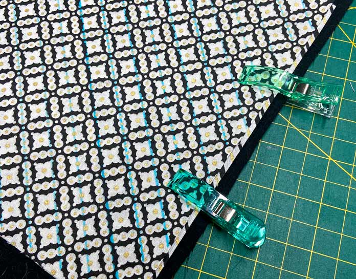 5 layers of black and white fabric are held with green clips and sit on top of a green cutting mat. The top fabric has blue diagonal lines drawn on it. Oliso M2Pro Mini Project Iron, FAIRFIELD Low-Loft Quilt Batting - Crib - 114 x 152cm (45″ x 60″), UNIQUE QUILTING Clever Clips, OLFA CHN-1 - Chenille / Textile Cutter, UNIQUE CREATIV Trim Pack - Black & White, FABRIC CREATIONS Burlap Fabric - Oyster - 45 x 61cm (18″ x 24″), FABRIC CREATIONS Faux Fur Fabric - Zebra - 45 x 76cm (18″ x 30″), UNIQUE SEWING Double Adjust Parachute Buckle - Plastic - 25mm (1″) White, CRAFTING ESSENTIALS Bottle of Buttons - Pink Tones - 75g (2.6oz), UNIQUE SEWING Invisible Magnetic Snaps - Sew-On - 12mm (1⁄2″) - 2 sets, UNIQUE QUILTING Stitch-N-Steam - 157 x 45cm (62″ x 18″), FABRIC CREATIONS Fabric Bundle (5pcs) - Metallic Gold - 45 x 53cm (18in x 21in), Oliso Pro TG1600 Pro Plus Smart Iron, UNIQUE QUILTING Clever Clips Large - 12pcs, UNIQUE SEWING 2-in-1 Dual-Tip Wash-out/Air Erasable Marking Pen - Fine, OMNIGRID Ruler - 121⁄2″ x 121⁄2″ (31.75 x 31.75cm), sewing tutorial