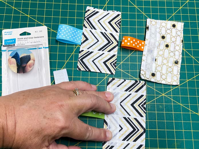 A hand presses down on a strip of hook and loop tape placed onto the back of a white, gold, and black rectangle of fabric. Two other rectangles of fabric with attached ribbons and the package from the hook and loop tape all sit on the green cutting mat. OLFA CHN-1 - Chenille / Textile Cutter, UNIQUE QUILTING Stitch-N-Steam - 157 x 45cm (62″ x 18″), FABRIC CREATIONS Fabric Bundle (5pcs) - Metallic Gold - 45 x 53cm (18in x 21in), UNIQUE SEWING Self-Gripping Fastener Strip - 25cm (10″) - White, UNIQUE Webbing 25mm x 10m (1″ x 11yd) - White, an ELAN Trench Buckle - 45mm (13⁄4″) - Brown, UNIQUE CREATIV Trim Pack - Black & White, UNIQUE QUILTING Clever Clips Small - 12 pcs, CLOVER 7807 - Hold it Precision Stiletto, UNIQUE SEWING Sewing Tools Holder, UNIQUE QUILTING Wool Pressing Mat - 18″ x 24″ - Grey, OLISO M2Pro Mini Project Iron with Solemate, UNIQUE QUILTING Clever Clips Large - 12pcs, UNIQUE SEWING Invisible Magnetic Snaps - Sew-On - 12mm (1⁄2″) - 2 sets, COSTUMAKERS Activewear One Way Separating Zipper 23cm (9″) - White - 1760, UNIQUE SEWING Double Adjust Parachute Buckle - Plastic - 25mm (1″) - White, free tutorial