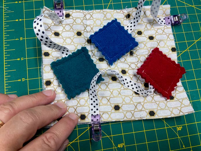 A hand holds down the edge of a white, gold, and black quilt sandwich. White ribbons with black polka dots have green, red, and blue wool felt squares attached to their ends, and are clipped to the edge of the quilt sandwich.  A green cutting mat is shown in the background. UNIQUE SEWING Self-Gripping Fastener Strip - 25cm (10″) - White, UNIQUE Webbing 25mm x 10m (1″ x 11yd) - White, ELAN Trench Buckle - 45mm (13⁄4″) - Brown, UNIQUE CREATIV Trim Pack - Black & White, UNIQUE SEWING Invisible Magnetic Snaps - Sew-On - 12mm (1⁄2″) - 2 sets,  SOFTKUT Pinking Shears (Scissors) - 81⁄2″ (21.6cm), COSTUMAKERS General Purpose Closed End Zipper 20cm (8″) - White - 1700, UNIQUE CREATIV Trim Pack - Red & Green, OLFA RTY-2/DX - Deluxe Ergonomic Handle Rotary Cutter 45mm, UNIQUE QUILTING Clever Clips Small - 12 pcs, UNIQUE SEWING Double Adjust Parachute Buckle - Plastic - 25mm (1″) - White, CRAFTING ESSENTIALS Bottle of Buttons - Pink Tones - 75g (2.6oz), UNIQUE Braided Elastic 3mm x 3.7m - White, free tutorial