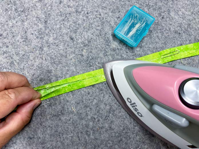 A hand is shown folding the lengthwise edges of a green strip of fabric into the middle while a small pink iron presses it into place. A fork pin holds the strip to a grey wool pressing mat. UNIQUE SEWING Invisible Magnetic Snaps - Sew-On - 12mm (1⁄2″) - 2 sets, COSTUMAKERS General Purpose Closed End Zipper 20cm (8″) - White - 1700, UNIQUE SEWING Double Adjust Parachute Buckle - Plastic - 25mm (1″) – White, UNIQUE QUILTING Clever Clips Small - 12 pcs, CLOVER 7812 Roll & Press, UNIQUE QUILTING Wool Pressing Mat - 18″ x 24″ - Grey, OLISO M2Pro Mini Project Iron with Solemate, CLOVER 240 - Fork Pins - .56mm, CLOVER 464/18 - Bias Tape Maker - 18mm (3⁄4″), Odif 505 Temporary Quilt Basting Adhesive Fabric Spray - 156g, FAIRFIELD Low-Loft® Quilt Batting - Crib - 114 x 152cm (45″ x 60″), free tutorial