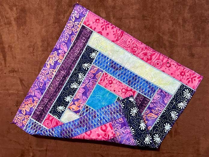 A square log cabin quilted block created with multicolored batik fabrics, using a flatlock serger stitch, showing one side with ribbon woven through the ladders of the stitch and the loopers showing the purple embroidery thread on the other side; PFAFF admire air 7000 serger 