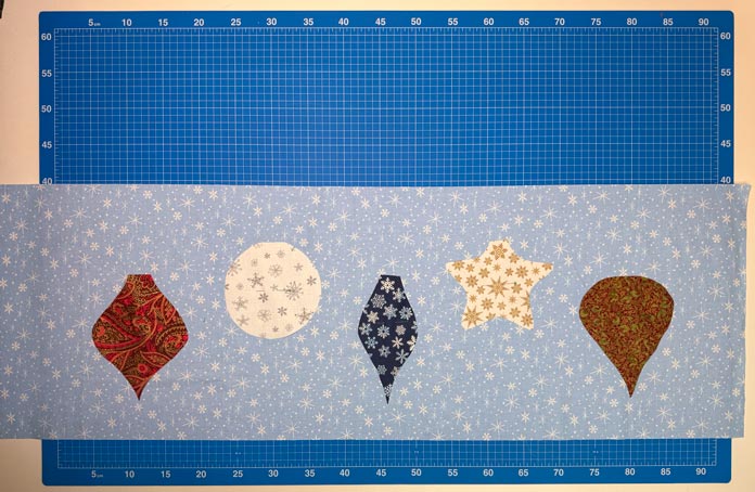 5 Christmas ornament shapes of various Christmas-themed fabrics placed on a light blue, snowflake print background fabric; Gütermann Dekor Metallic Thread 200m – Gold, Gütermann Dekor Metallic Thread 200m - Col. 41, Gütermann Dekor Metallic Thread 200m – Red, Gütermann Dekor Metallic Thread 200m - Christmas Green, Gütermann Dekor Metallic Thread 200m - Col. 1005, Gütermann Denim Thread 100m – Black, UNIQUE Baby Rick Rack 3mm x 3.7m – Scarlet, UNIQUE Medium Rick Rack 6mm x 2.3m - Emerald Green, UNIQUE Jumbo Rick Rack 10mm x 2.3m - Metallic Gold, SCHMETZ #1743 Metallic Needles Carded - 80/12 - 5 count, SCHMETZ #1752 Metallic Needles Carded - 90/14 - 5 count, LDH 8½“ Craft Scissors – Purple, HeatnBond Lite Iron-On Adhesive - 43cm x 30m (17″ x 32 ¾yds) bolt, HeatnBond Stitch n Sew Tear-Away Stabilizer - 50.8cm x 22.8m (20″ x 25yds), oliso M3Pro Project Iron – Coral, OLFA 45mm RTY-2/DX/MAG Ergonomic Rotary Cutter – Magenta, UNIQUE Quilting Wool Pressing Mat - 18″ x 24″ - Grey