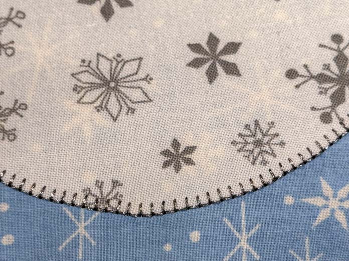 A double blanket stitch of silver metallic thread on the edge of a white and grey fabric stitched to a light blue snowflake print background fabric; Gütermann Dekor Metallic Thread 200m – Gold, Gütermann Dekor Metallic Thread 200m - Col. 41, Gütermann Dekor Metallic Thread 200m – Red, Gütermann Dekor Metallic Thread 200m - Christmas Green, Gütermann Dekor Metallic Thread 200m - Col. 1005, Gütermann Denim Thread 100m – Black, UNIQUE Baby Rick Rack 3mm x 3.7m – Scarlet, UNIQUE Medium Rick Rack 6mm x 2.3m - Emerald Green, UNIQUE Jumbo Rick Rack 10mm x 2.3m - Metallic Gold, SCHMETZ #1743 Metallic Needles Carded - 80/12 - 5 count, SCHMETZ #1752 Metallic Needles Carded - 90/14 - 5 count, LDH 8½“ Craft Scissors – Purple, HeatnBond Lite Iron-On Adhesive - 43cm x 30m (17″ x 32 ¾yds) bolt, HeatnBond Stitch n Sew Tear-Away Stabilizer - 50.8cm x 22.8m (20″ x 25yds), oliso M3Pro Project Iron – Coral, OLFA 45mm RTY-2/DX/MAG Ergonomic Rotary Cutter – Magenta, UNIQUE Quilting Wool Pressing Mat - 18″ x 24″ - Grey