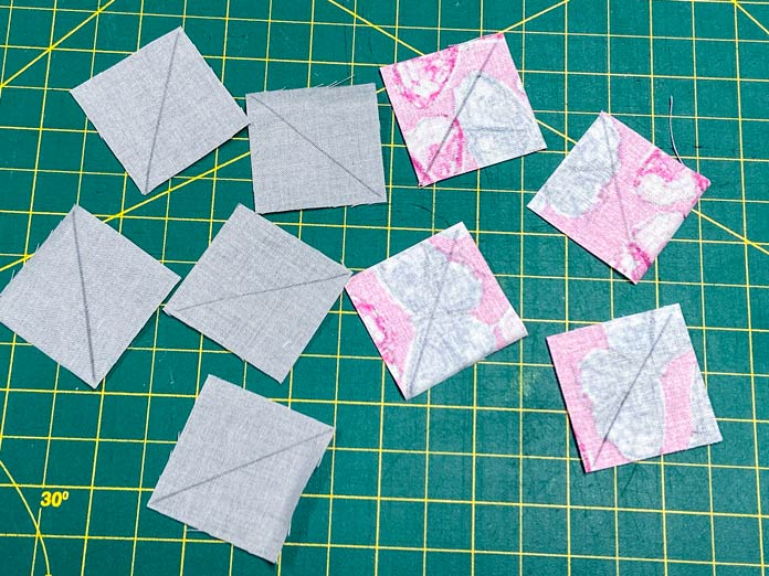 Five gray squares of fabric and four pink squares of fabric are shown laying on a green cutting mat with their right sides down. A diagonal line has been drawn on the back of each of them.