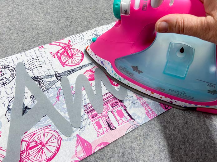 A pink iron is being used to iron a fusible applique design to the front of a gray, black, white and pink piece of fabric. A mottled gray wool pressing mat is in the background.