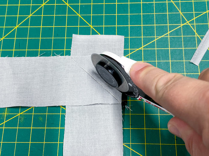 A hand uses a white rotary cutter to trim away the excess fabric from two gray fabrics that have been sewn together with a diagonal seam. A green cutting mat is in the background. 