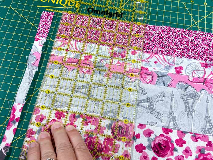 A hand is shown holding a rotary ruler with a yellow grid on top of a pink, white and gray strip set of fabric. The left hand edge of the strip set has been cut off to square up the fabrics. Everything sits on top of a green cutting mat. 