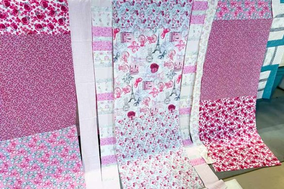 Pink and white fabrics that have been sewn together into rows hang on the rail of a longarm sewing machine.