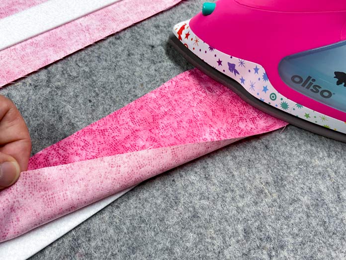 A hand folds back the corner of a strip of pink fabric to show the white strip of interfacing underneath. A pink iron is ironing one end of the strip on top of a grey pressing mat.