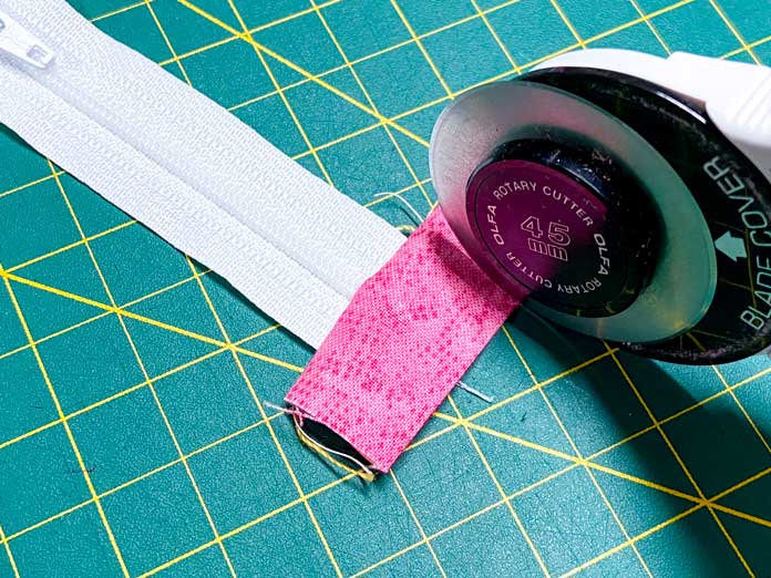 A hand uses a white rotary cutter to trim away excess pink fabric from the side of a white zipper. A green cutting mat is shown in the background.