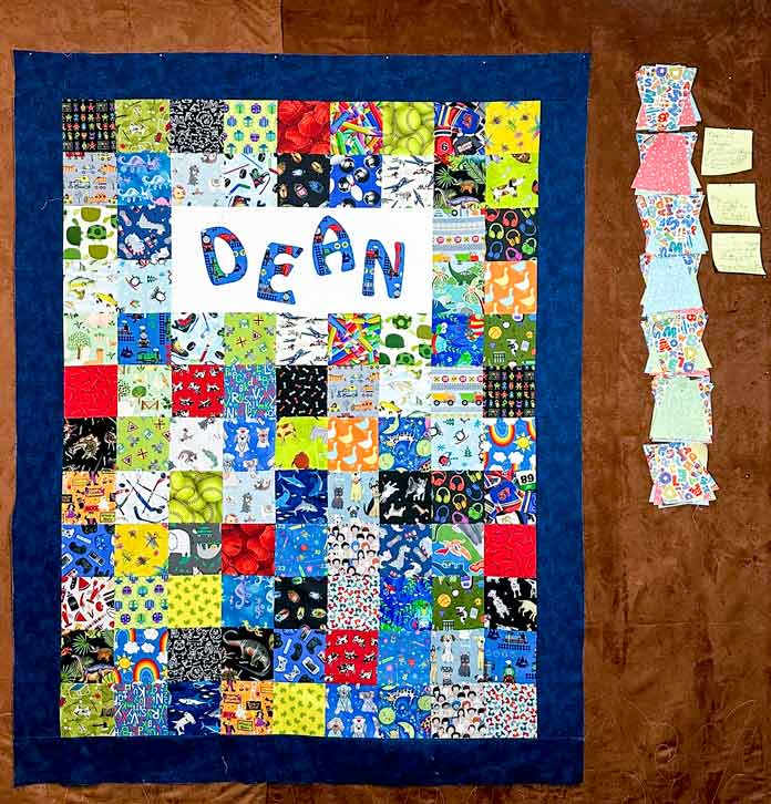 5” squares of unique kid prints, stitched together in 12 rows of 9 block. The 3rd and 4th row have a 2X5 block section of white with an embroidered blue applique letters forming the name “DEAN”. and the whole thing has a navy 4.5” border around it.