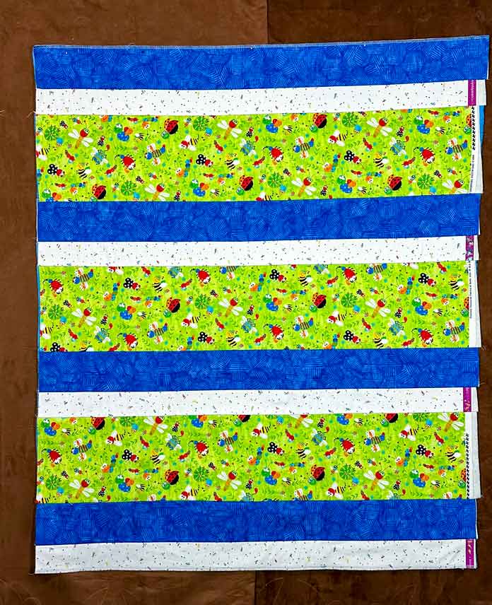 Strip quilt (from bottom edge) in white tiny print 2.5” strip, blue batik 4” strip, and green bug print 8.5” strip, repeated 3X, ending with a white and blue strip. All selvedges on the right uneven edge showing.