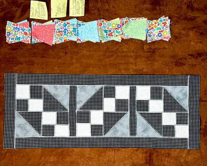 A runner with 3 blocks Black, gray and white silver metallic fabrics in a “Road to Oklahoma” pattern with black/silver sashing and borders. All the seams are exposed, with decorative gray/silver metallic thread.