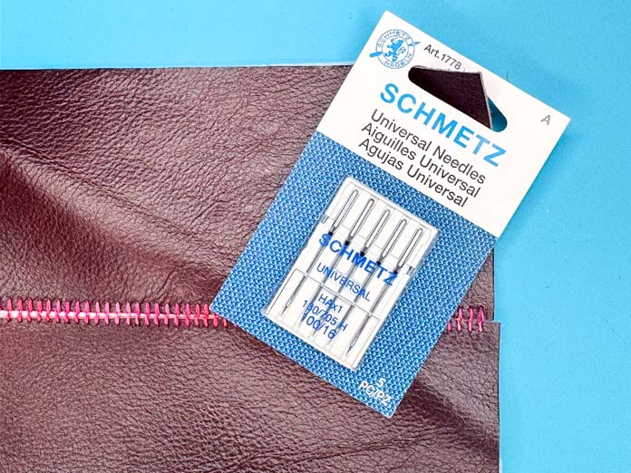 A package of SCHMETZ 100/16 Universal sewing machine needles on a piece of leather.