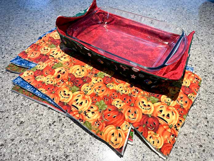 Thanksgiving pumpkin fabric 9” x 13” casserole cozy with a glass casserole inside on top of 3 assorted holiday fabric kits (fabric and batting) ready to sew up more.
