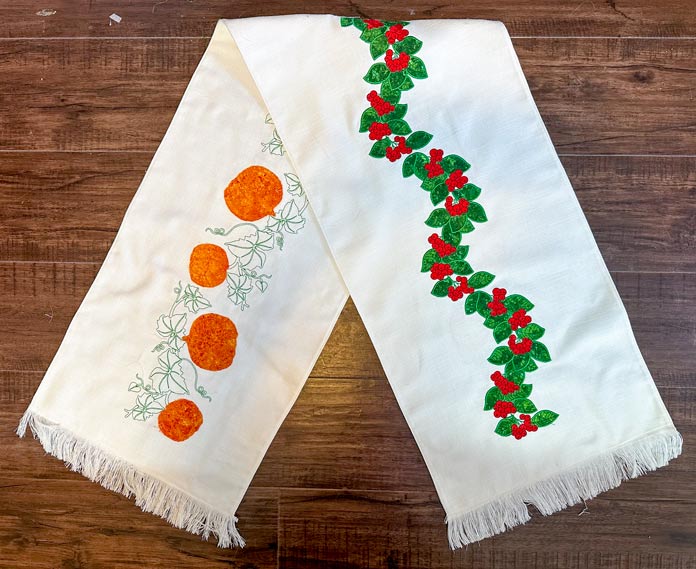 A cream reversible runner 15” x 67”, embroidered with orange applique pumpkins and green vines on one side and Appliqued Green Holly leaves and Red Berries on the other side. Both ends are frayed for a fringe effect.