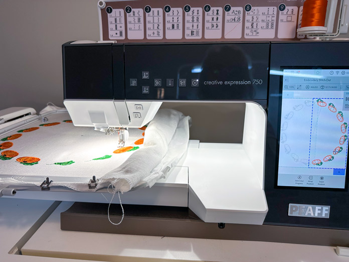 A picture of a deep charcoal and white sewing /embroidery machine with a large computer screen, turned on waiting to be used. A white embroidery arm attached, and a large 360X350 hoop attached with a cream piece of fabric inserted