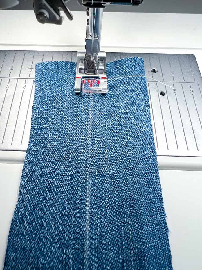 Close-up of the PFAFF Creative Icon 2 sewing machine with Fancy Stitch Foot 2A attached, aligned on denim fabric. A fine chalk or water-soluble marker line has been drawn to guide the stitching path, helping to keep the decorative ribbon stitches straight. The feed dogs and throat plate markings are clearly visible, providing additional support for precision. This step sets up the base stitch path before the ribbon is manually added and stitched down in controlled segments using the Start/Stop button instead of the foot pedal.