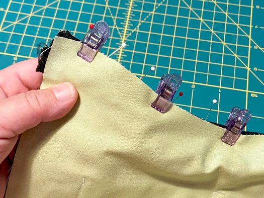 A hand holds a green piece of lining clipped to a zipper and a second piece of fabric. Pins can also be seen along this top edge, and a green cutting mat is in thebackground.