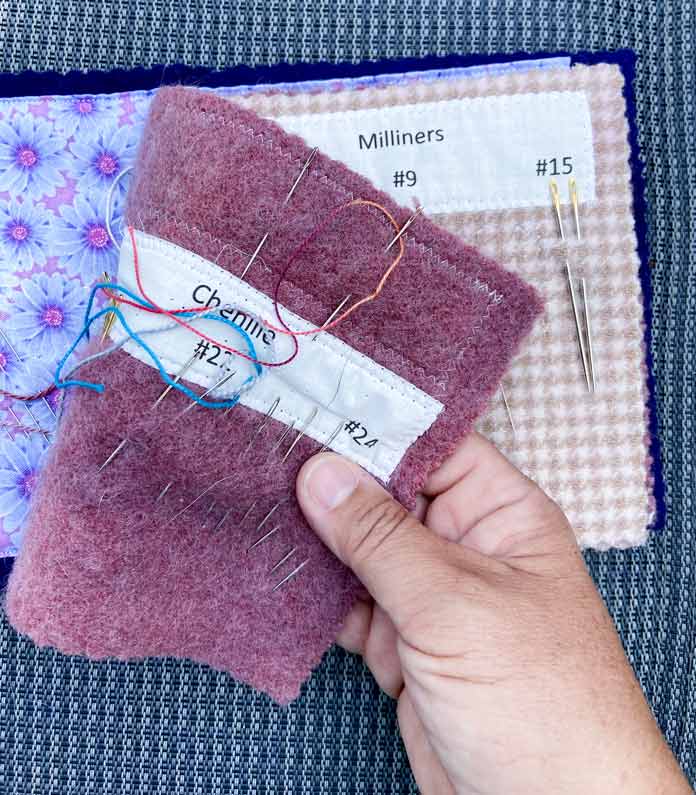 A hand is shown holding one of the pages of the pink and purple needle book. There are labels in the book and needles are stuck into each page. Some of the needles are threaded with colorful threads; DMC Embroidery Floss, DMC Cotton Perle Thread, Clover Gold Eye Chenille Needles (No. 22)