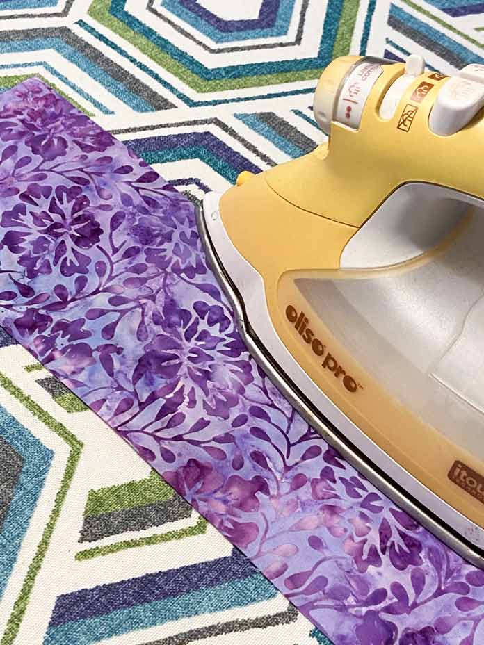A yellow Oliso Pro iron is shown pressing a strip of purple floral batik fabric in half on top of a white, teal, blue and grey geometric ironing board.