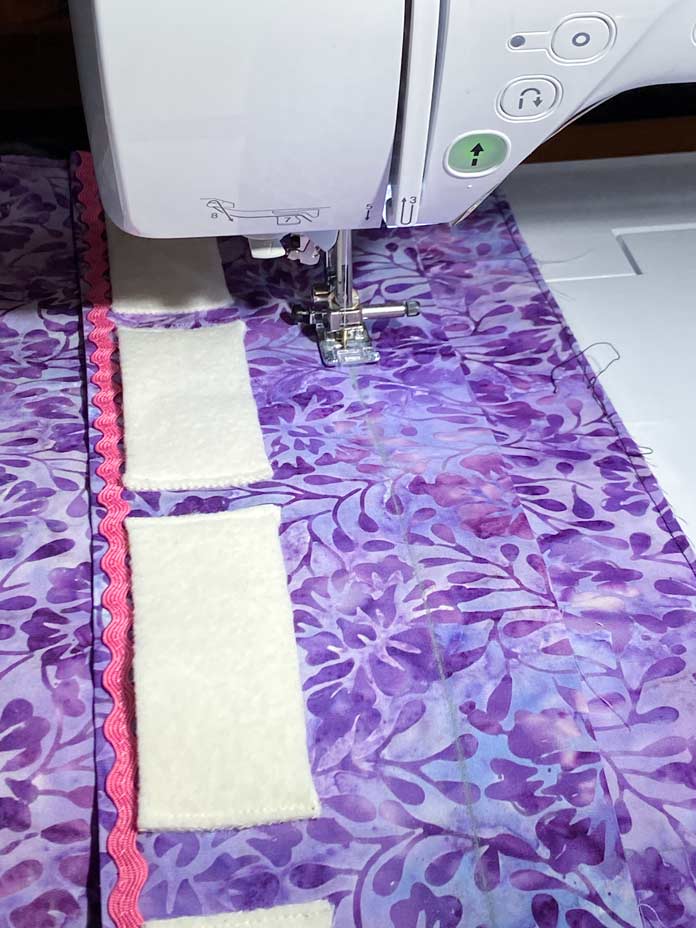 Purple fabric with white rectangles of wool lined up beside pink rick rack on top, sitting on the bed of a sewing machine. Brother NQ900 sewing machine, UNIQUE quilting Clever Clips, UNIQUE Medium Rick Rack