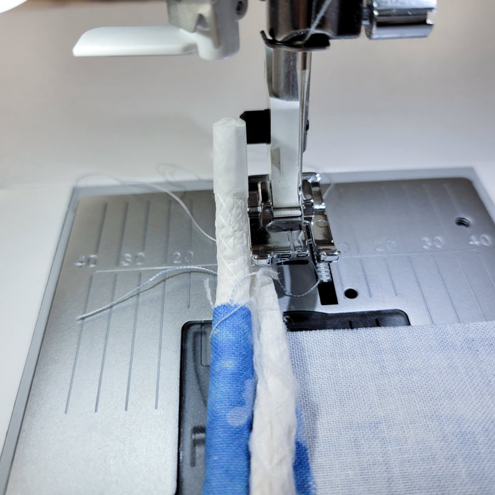 Two lengths of white cord with one wrapped in a blue fabric in front of a metal presser foot on a sewing machine; Husqvarna Viking Opal 670