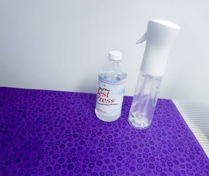 Mary Ellen’s Best Press refill bottle and spray bottle sitting on pressed purple fabric with no wrinkles.