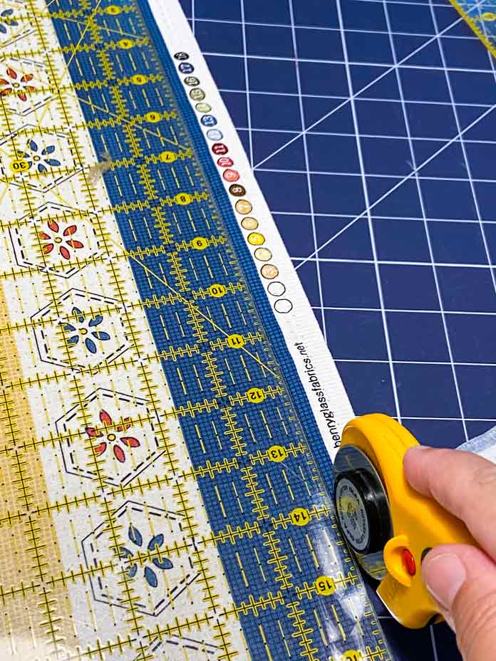 A hand is shown holding a yellow rotary cutter to trim away excess fabric from the panel using a quilting ruler. A blue rotary cutting mat is shown in the background; Omnigrid ruler - 6" x 24", OLFA Rotary Cutter, OLFA Double Sided Cutting Mat (navy blue)
