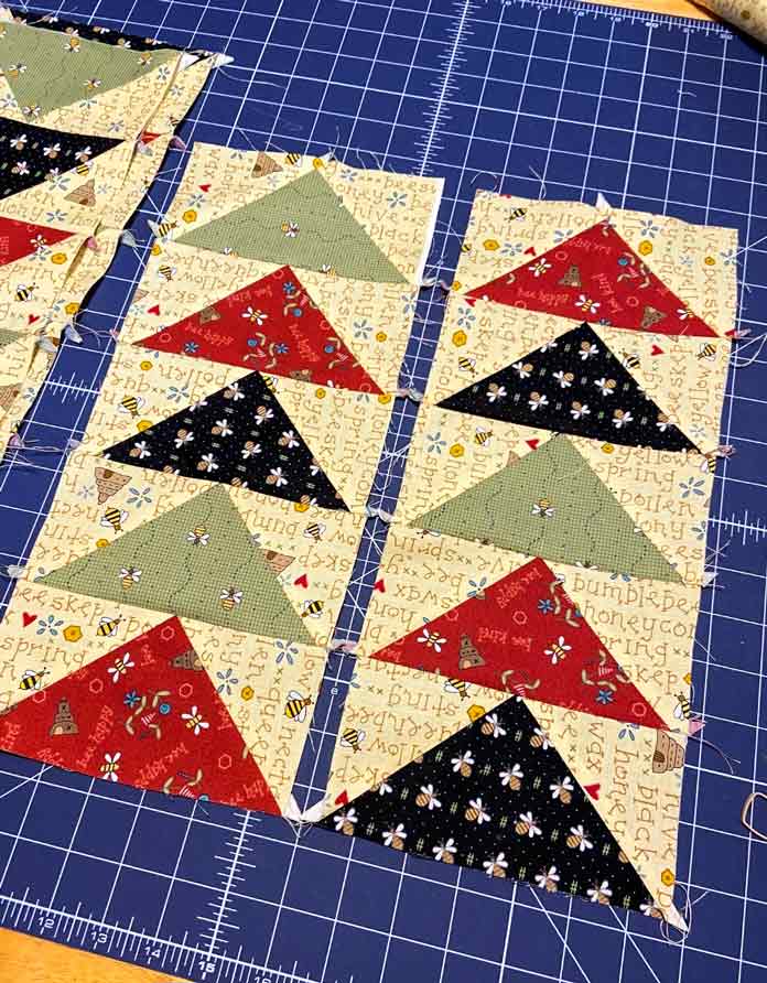 Two rows of five flying geese blocks in red, black green and yellow lay on a dark blue cutting mat; OLFA Double Sided Cutting Mat (navy blue)