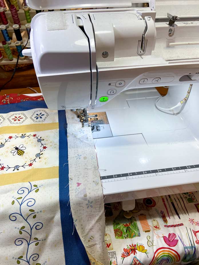 A narrow yellow border being sewn to a bee theme panel is shown laying on the bed of a white and blue sewing machine. The sew button on the sewing machine is glowing green; Brother NQ900