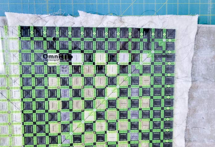 A large square quilting ruler on a black, gray, and gold table runner, quilted but not trimmed